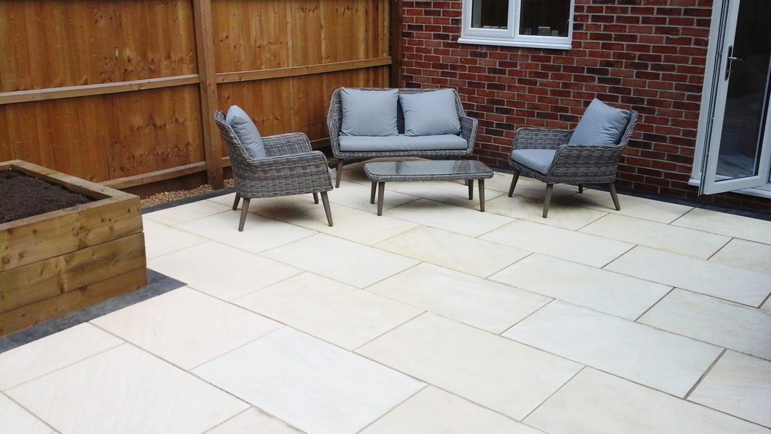 A spacious outdoor patio area featuring three grey cushioned wicker chairs and a matching glass-top table. The patio is paved with light-colored tiles and bordered by a wooden fence and a wooden raised garden bed.