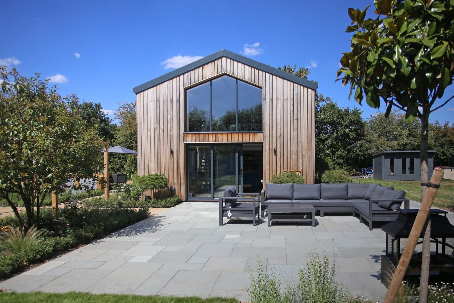 Modern wood-clad house with large central windows, surrounded by trees and plants. Spacious patio features gray stone tiles, an L-shaped outdoor sofa, a table, and chairs. Clear blue sky overhead.