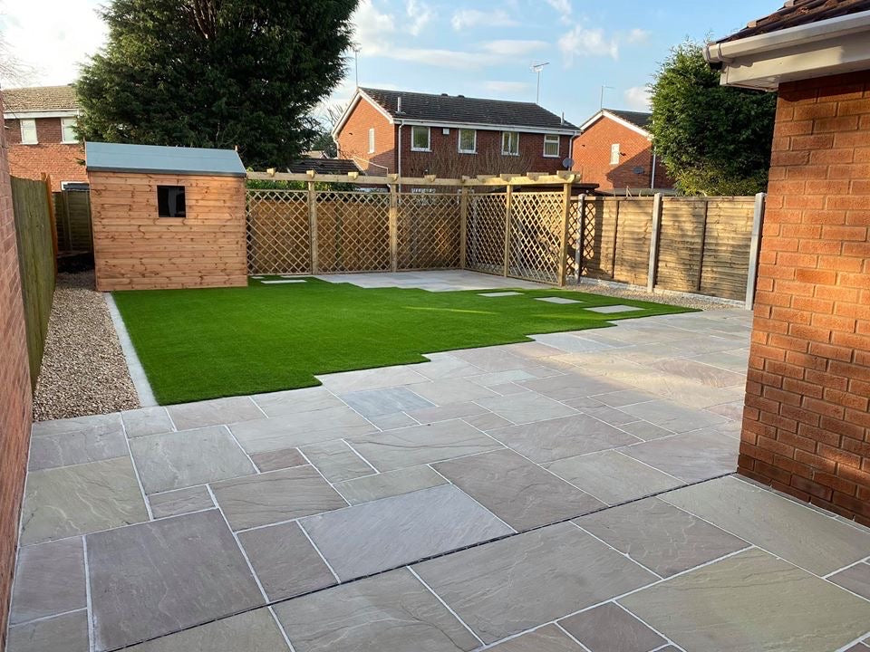 A backyard with a patio made of stone tiles, adjacent to a patch of artificial grass. There is a wooden shed in the corner, wooden fencing, and a few brick houses in the background under a clear sky.