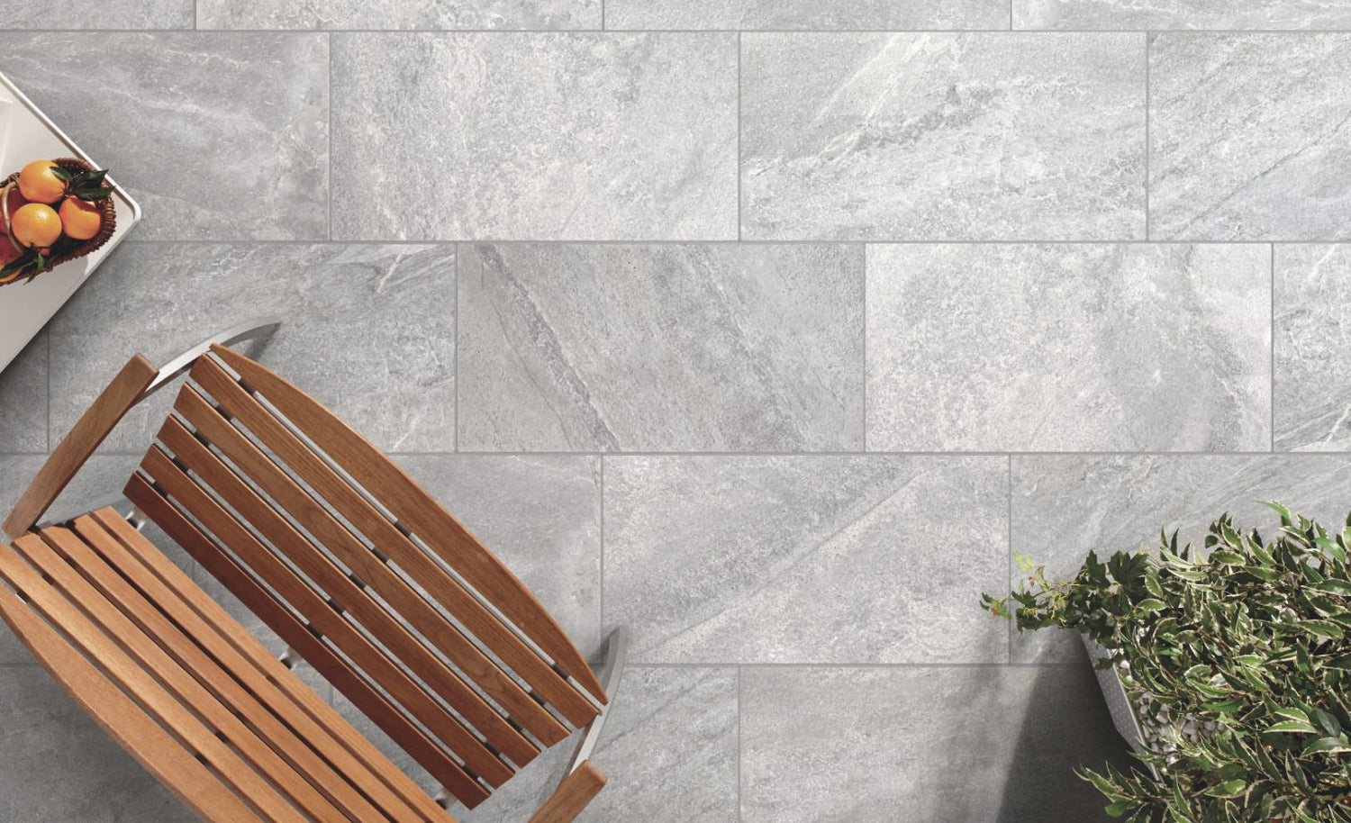 Top view of a grey stone tile floor with a brown wooden bench on the left, a fruit basket on the top left corner, and a plant in the bottom right corner.