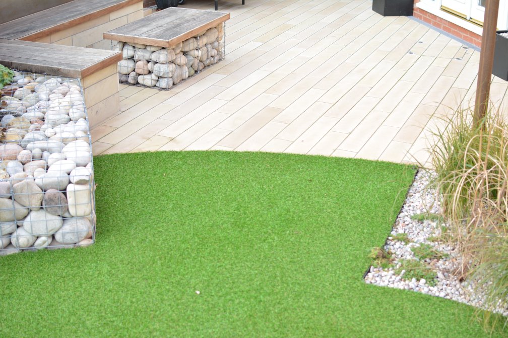 Outdoor seating area with a wooden deck and bench, featuring stones encased in wire. Adjacent to the deck is a patch of artificial grass bordered by white pebbles and a few plants.