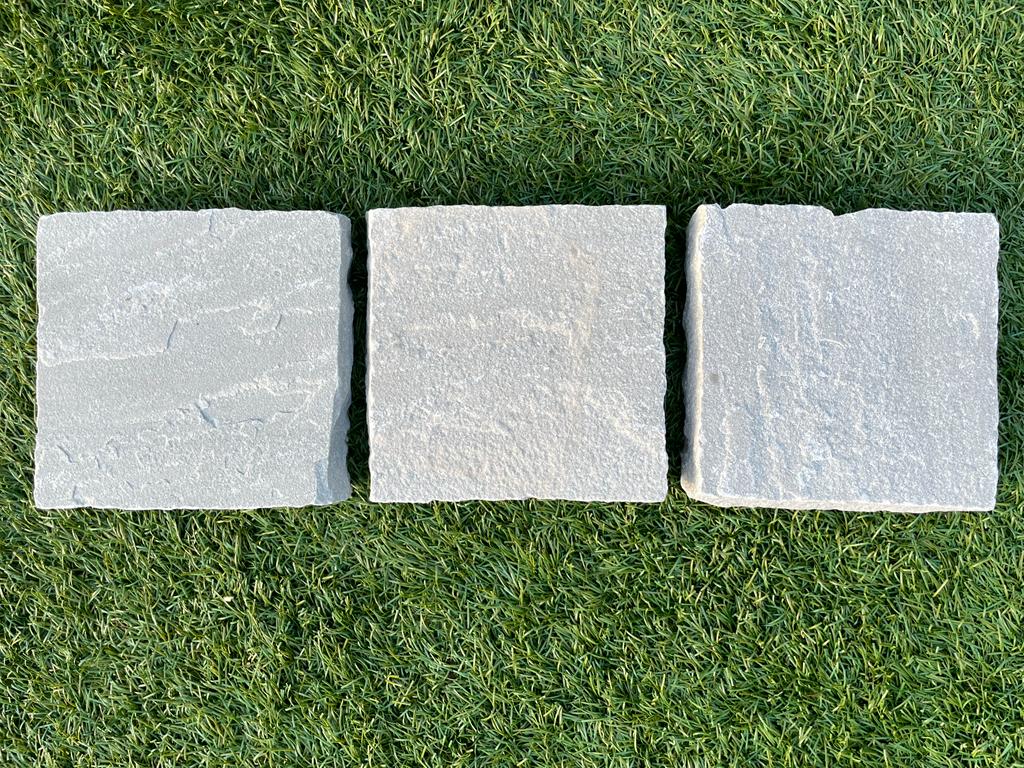 Three square stone pavers arranged in a row on a patch of green grass. The pavers have a rough texture and slightly varied shades of gray.