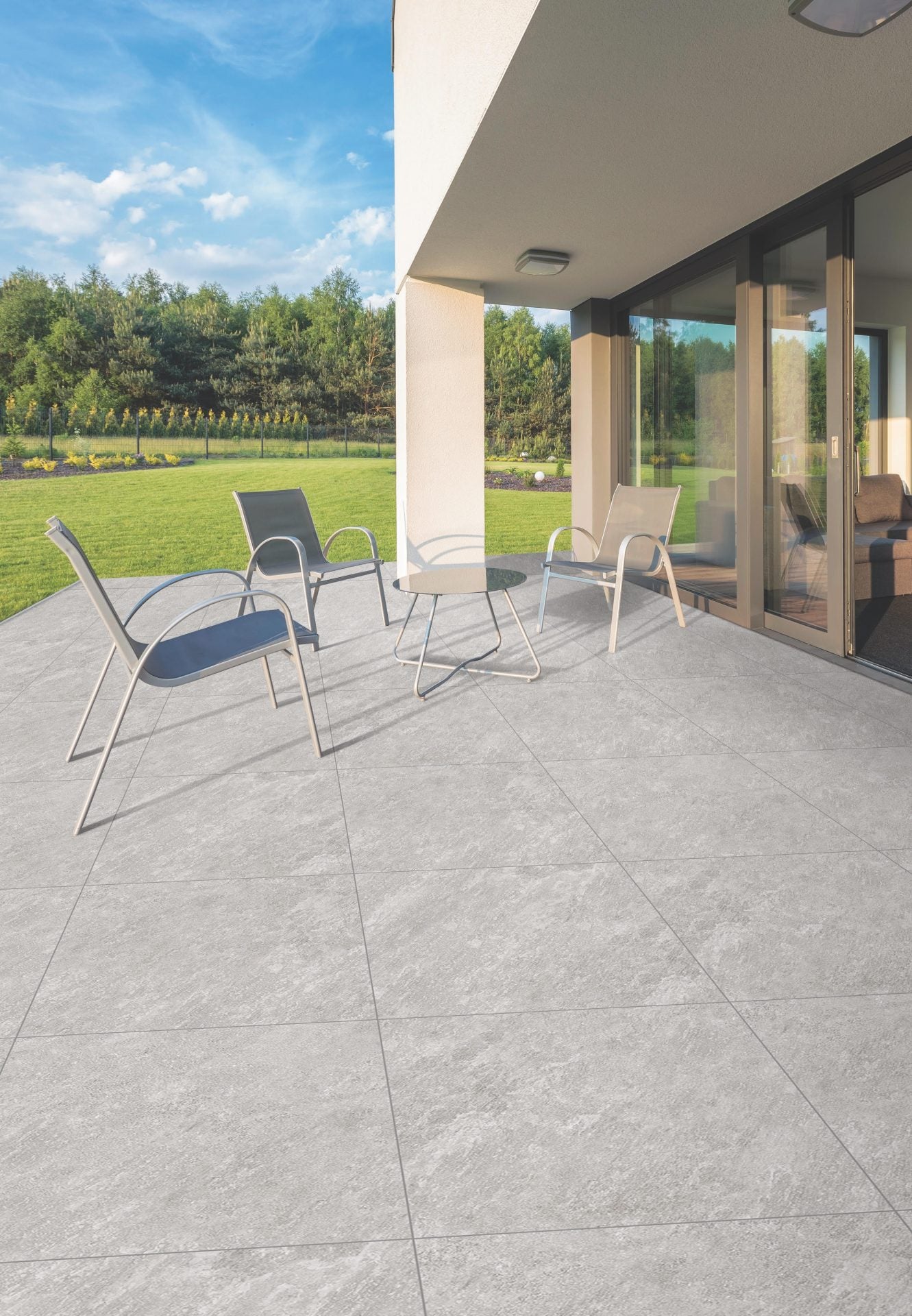 Modern outdoor patio with light gray tiles, featuring three minimalist chairs and a round glass table. Patio is adjacent to a glass door, overlooking a green lawn and a forested area under a clear blue sky.