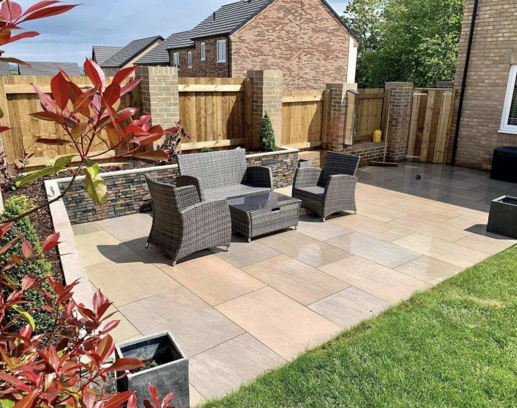 A neatly paved backyard patio with gray wicker furniture, including two chairs and a small table. It's surrounded by a wooden fence and bordered by a well-maintained garden with a few plants and shrubs.