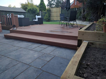 A backyard deck with wooden steps, durable Paving Shopper Kota Black Limestone Paving, patio furniture including a round table with two chairs, and a surrounding landscaped garden area.