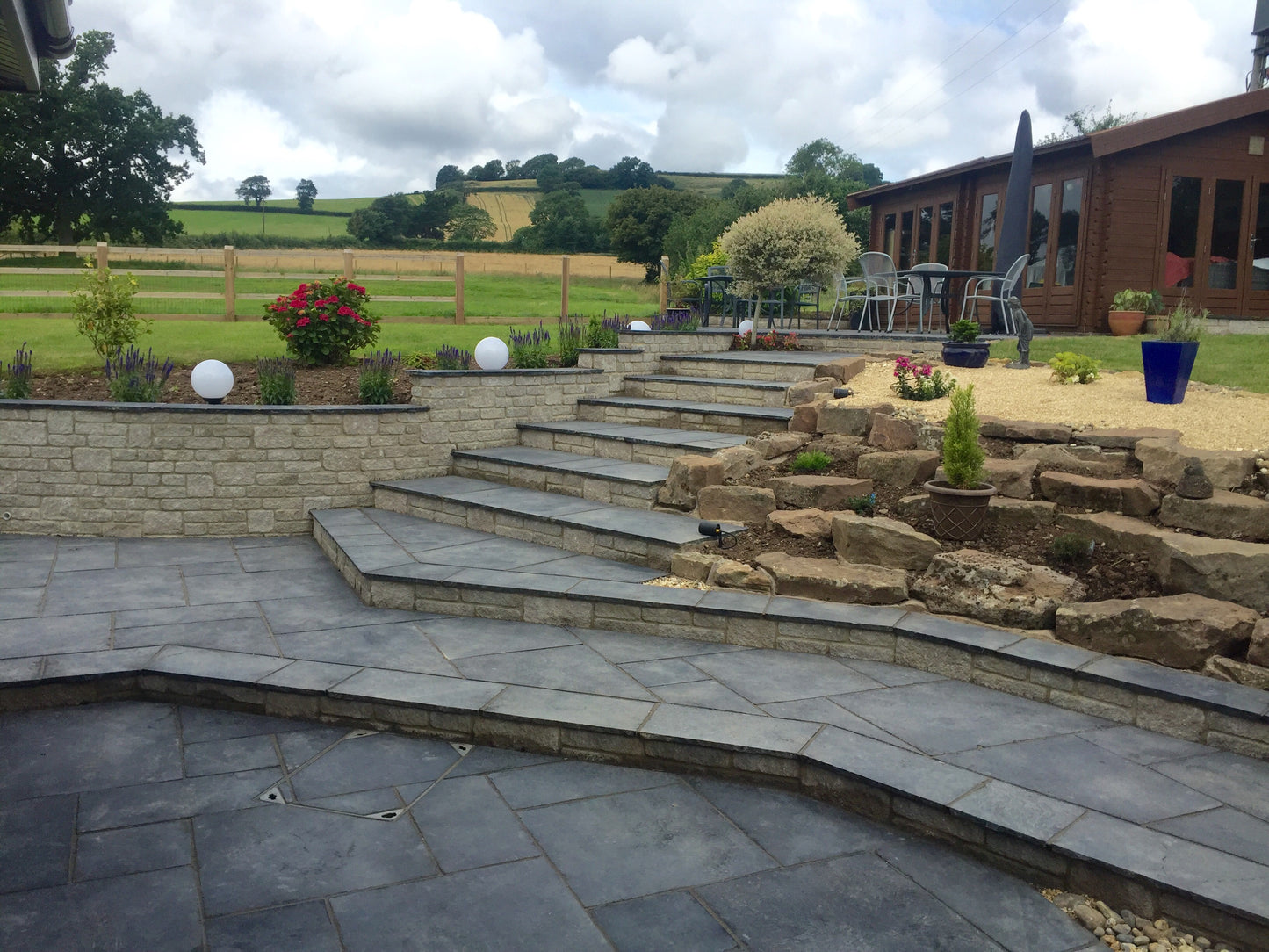 A landscaped backyard features a stone patio with steps made of durable, slip-resistant Paving Shopper's Kota Black Limestone Paving, alongside garden beds with decorative plants, outdoor furniture, and countryside views in the background.