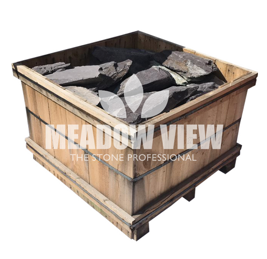 A wooden crate filled with large, irregularly shaped stones. The text "Paving Shopper - Blue Slate Rockery 250mm" is printed on the side of the crate.