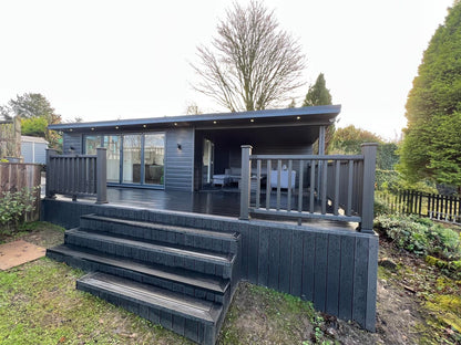 Chic cabin enveloped in a dark grey hue, boasting expansive windows and an elevated deck constructed with Paving Shopper's Composite Decking (58.24m2), featuring scratch-resistant railing and steps, nestled among lush trees and greenery.