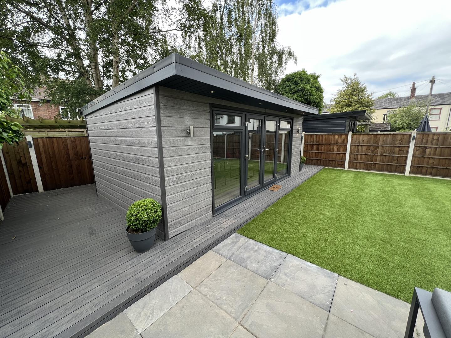 Modern garden shed with glass doors, surrounded by a Silver Salt Lake composite decking from Paving Shopper that spans 58.24m² and is resistant to scratches, complemented by artificial grass and enclosed by a durable wood grain fence.