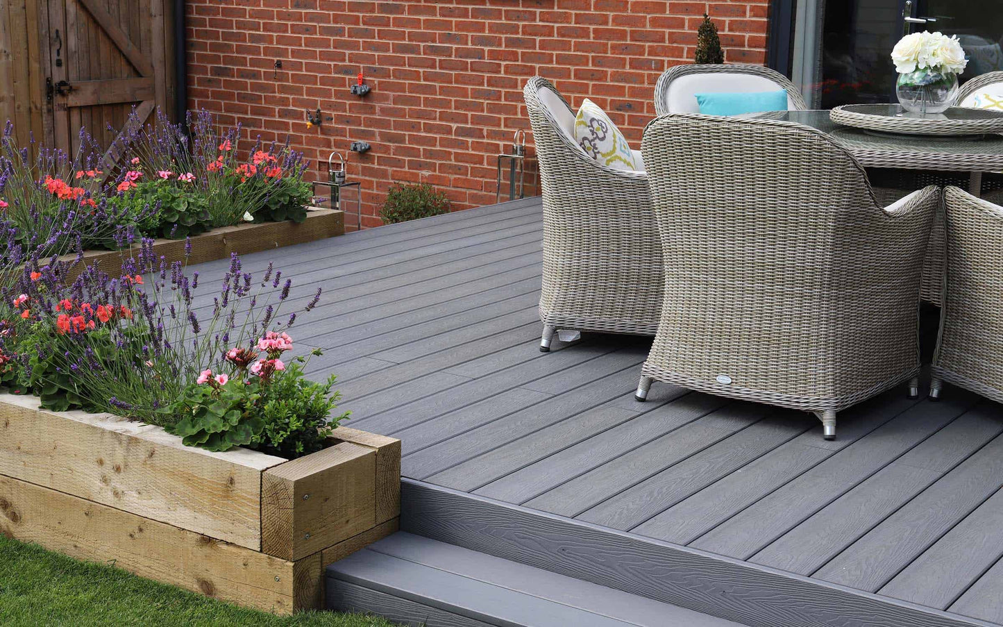 The Composite Decking - 58.24m2 in Dark Grey from Paving Shopper showcases a durable wood grain design, accompanied by wicker chairs and a round table near a red brick wall. On the left side, a wooden planter box filled with vibrant flowers provides an easy-to-manage option for enhancing your outdoor space.