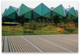 A futuristic building with a jagged, geometric glass facade showcases scratch-resistant surfaces and sits against a backdrop of greenery, featuring Paving Shopper's low-maintenance Composite Decking in Wild Field Brown, covering 58.24m² in the foreground.