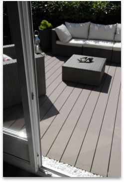 An inviting outdoor patio featuring wicker furniture with white cushions and a square coffee table made from durable wood grain. The space is accessible through a glass door and surrounded by vibrant greenery, all set on 58.24m2 of Dark Grey Composite Decking by Paving Shopper.